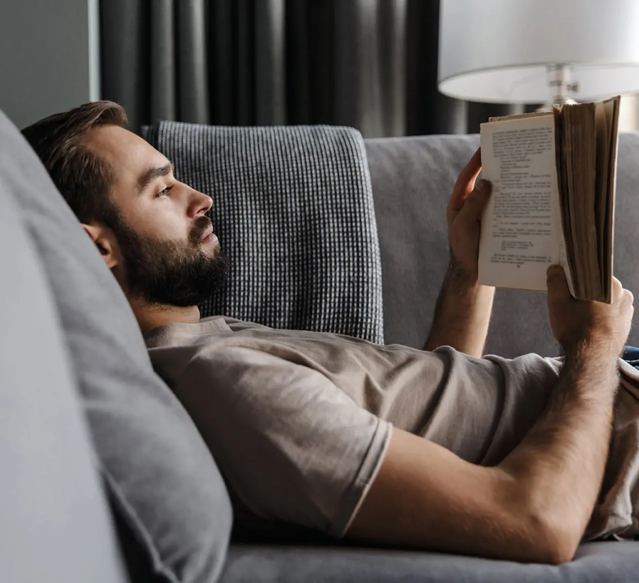 Mann liegt entspannt auf einem grauen Sofa und liest ein Buch. Gemütliche Wohnatmosphäre mit weicher Decke, warmem Licht und ruhigem Hintergrund – Symbol für Ruhe und Entspannung im Alltag.
